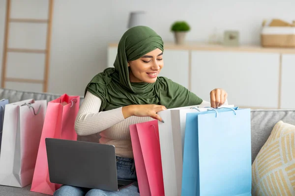 Mujer árabe joven en hijab sentado en el sofá con el ordenador portátil, mirando dentro de bolsas de regalo, comprobando entregado pedido en línea en casa —  Fotos de Stock