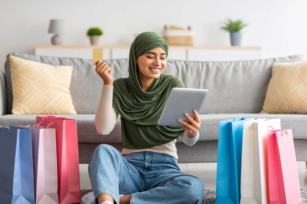 Encantadora joven árabe en hijab compras en línea desde la tableta PC, utilizando la tarjeta de crédito, sentado en el suelo con bolsas de regalo —  Fotos de Stock