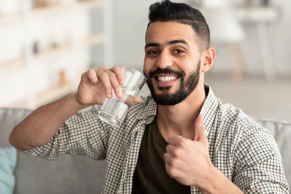 Fique hidratado. Sorrindo árabe cara bebendo água mineral de vidro e mostrando polegar acima gesto em casa — Fotografia de Stock