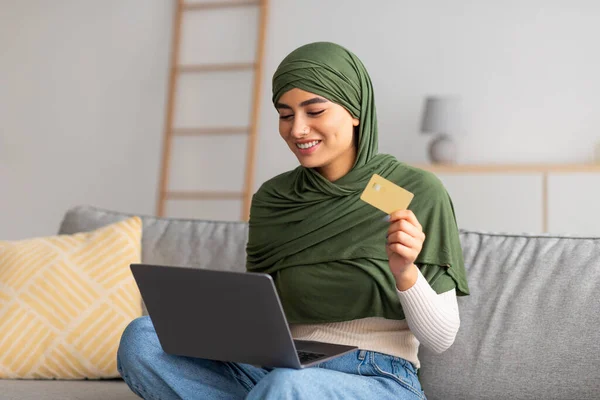 Genç Arap kadın tesettürde kredi kartı ve dizüstü bilgisayar kullanıyor, kanepede online alışveriş yapıyor, evden internet üzerinden mal alıyor. — Stok fotoğraf