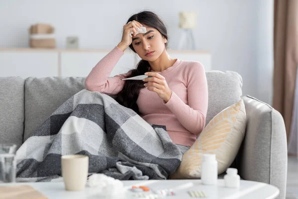 Désespéré jeune femme hindoue triste avec carreaux, souffrent de grippe, s'assoit sur le canapé et regarde thermomètre dans le salon — Photo