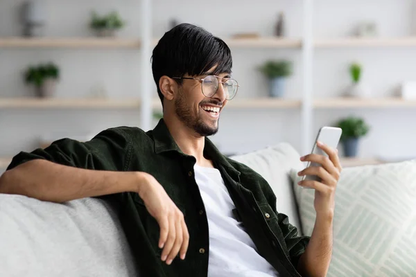 Щасливий індійський хлопець сидить на дивані, використовуючи смартфон — стокове фото