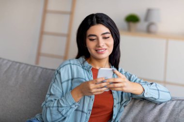 Güzel genç Arap bayan cep telefonu kullanıyor, online toplantı yapıyor, kanepede oturuyor, evdeki sosyal ağlara takılıyor.
