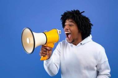 Harika haberlerim var. Telaşlı Afrikalı Amerikalı genç adam mavi stüdyo arka planında megafona bağırıyor.