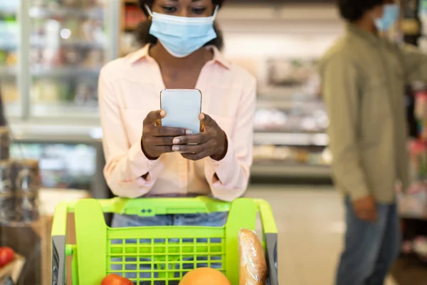 Μαύρη γυναίκα κάνει ψώνια χρησιμοποιώντας κινητό τηλέφωνο φορώντας μάσκα στο σούπερ μάρκετ — Φωτογραφία Αρχείου