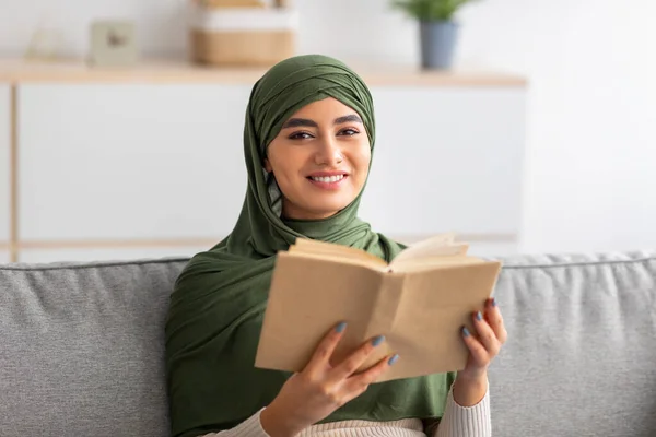 Tranquilos pasatiempos en casa. Mujer árabe milenaria en el libro de lectura de hijab, sentada en el sofá en la sala de estar — Foto de Stock