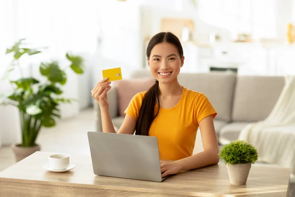 Cheerful asian lady with credit card using laptop at home, ordering food or clothes online, sitting at desk — Stock Photo, Image