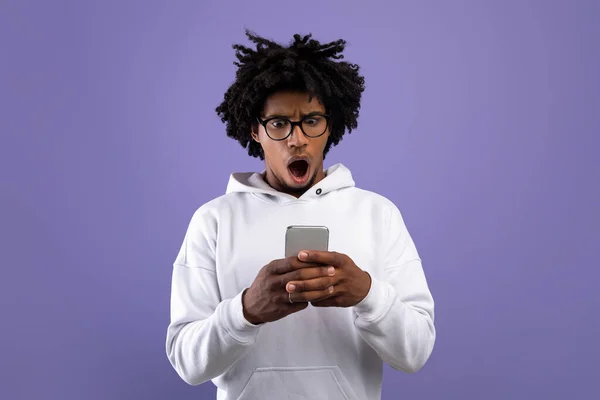 Frustrované černošky teen chlap zírá na smartphone v šoku přes fialové studio pozadí — Stock fotografie