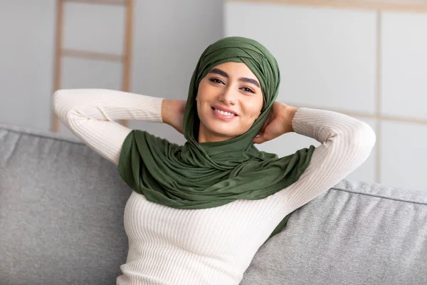 Улыбающаяся арабка тысячелетия в хиджабе, отдыхающая на диване с руками за головой, отдыхающая дома — стоковое фото