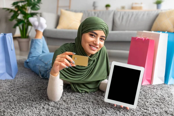 Joven árabe en hijab tumbada en el suelo con tarjeta de crédito y tableta con pantalla vacía, rodeada de bolsas de regalo, maqueta —  Fotos de Stock