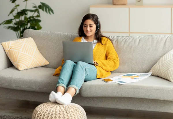 Концепция телекоммутации. Возбужденная арабская женщина, сидящая на диване и работающая онлайн на ноутбуке, в наушниках — стоковое фото