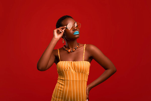 Glamorous african american young lady in bright summer outfit, sunglasses, accessories and makeup posing alone on red studio background, touching eyewear and looking at copy space
