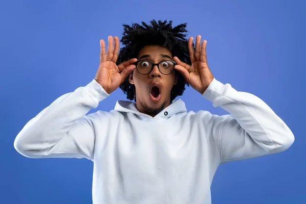 Emocionální černošky teenager otvírá ústa v šoku, dotýká brýle, dívá se na kameru na modrém pozadí studia — Stock fotografie
