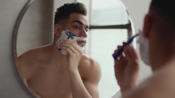 Self grooming concept. Close up portrait of young shirtless arab guy shaving his face, looking at mirror at bathroom — Stock Video