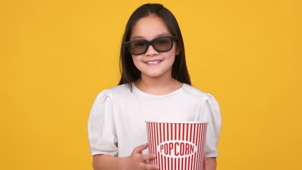 Dětská zábava. Studio portrét malé asijské dívky ve 3D brýlích drží popcorn kbelík a usmívá se na kameru — Stock video