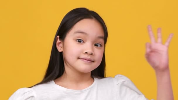 Разрешение ребенка. Студийный портрет милой маленькой азиатской девочки, показывающей ОК жест и улыбаясь в камеру, оранжевый фон — стоковое видео