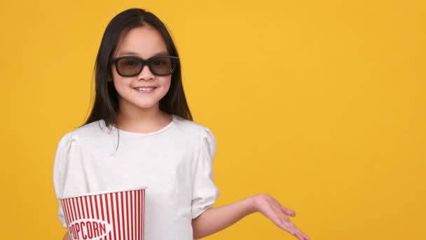 Щаслива маленька азіатська дівчинка в 3D окулярах з попкорн відро показує щось з простягнутою рукою, вільне місце — стокове відео