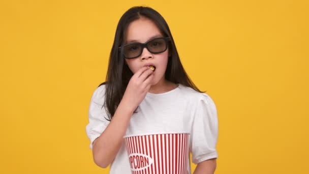 Hora del cine. Pequeña chica asiática con gafas 3d comiendo palomitas de maíz dulce de cubo y mirando a la cámara, disfrutando del cine — Vídeo de stock