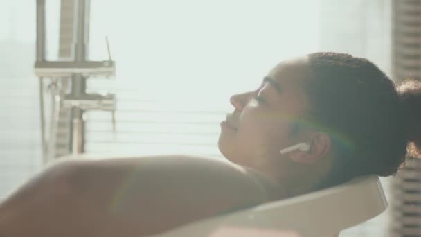 Banyoda rahatla. Genç Afro-Amerikan kadının banyoda kablosuz kulaklıkla müzik dinlemesini yakından çek. — Stok video