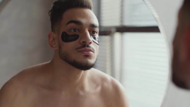 Мужская косметика. Молодой ухоженный араб с гелевыми пластырями под глазами, смотрящий в зеркало в ванной комнате — стоковое видео