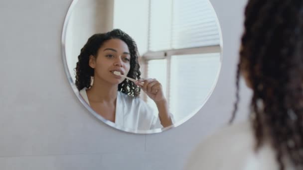 Tandheelkundige zorg concept. Over schouder uitzicht van jonge Afrikaanse Amerikaanse vrouw poetsen tanden, kijken naar spiegel op badkamer — Stockvideo