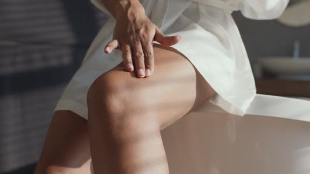 Cura del corpo. Giovane donna nera irriconoscibile che applica crema lifting anti cellulite sulle gambe, seduta in bagno — Video Stock