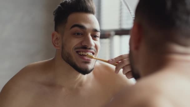 Higiene oral. Jovem homem árabe sem camisa positiva escovando os dentes com escova de dentes, olhando para o espelho no banheiro — Vídeo de Stock