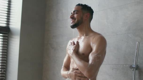 Mladý pozitivní svalnatý středovýchodní chlap se ráno sprchuje, stojí pod kapkami vody a myje si tělo — Stock video