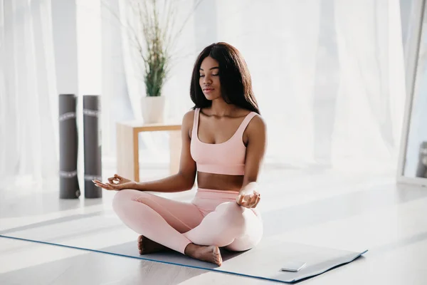 Jovem mulher negra pacífica sentada em pose de lótus, meditando, alcançando a paz interior e harmonia no tapete em casa — Fotografia de Stock