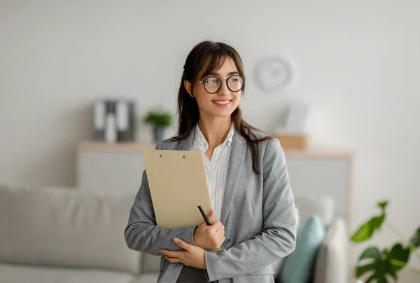 Porträt einer arabischen Psychotherapeutin mit Klemmbrett, die zur Seite schaut und lächelt, im modernen Büro steht, freier Raum — Stockfoto