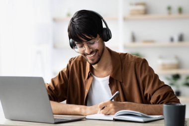 Neşeli bir ortadoğulu, web sitesine katılıyor, dizüstü bilgisayar kullanıyor, not alıyor.
