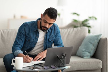 Uzak iş görüşmeleri sırasında not alan, bilgisayarını kullanan, uzaktan çalışan genç bir Arap.