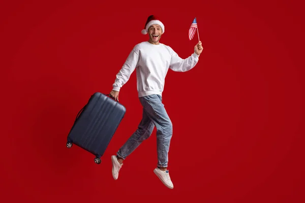 Podekscytowany młodzieniec w Santa Hat skacze z amerykańską flagą i walizką — Zdjęcie stockowe
