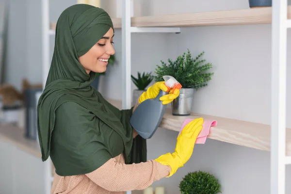 Μουσουλμάνα νεαρή γυναίκα καθαρίζει ράφια με απορρυπαντικό στο σπίτι — Φωτογραφία Αρχείου