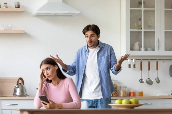 不快乐的年轻欧洲女人拿着智能手机，在厨房里对生气的尖叫男人视而不见 — 图库照片