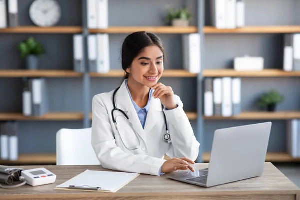 Sorrindo jovem indiano médico feminino em casaco branco trabalha no laptop no local de trabalho no escritório da clínica interior — Fotografia de Stock