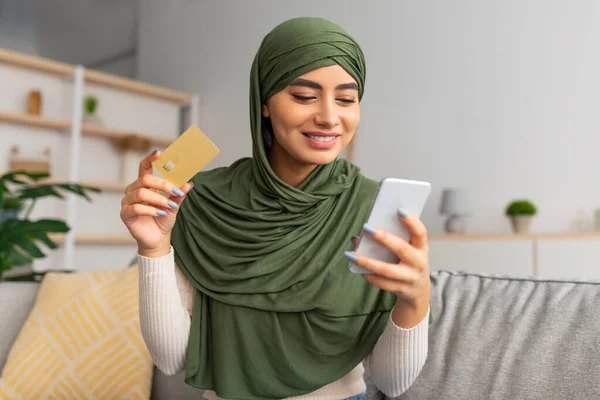 快乐的阿拉伯女人，头戴头巾，坐在沙发上，拿着智能手机和信用卡，在家里上网购物 — 图库照片