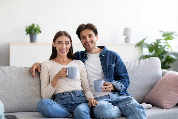 Mosolygó vidám millenniumi európai férfi és nő ül a kanapén egy csésze forró ital, élvezze a szabadidőt — Stock Fotó