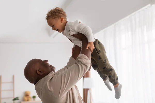 Africano avô segurando pequeno neto no ar em casa — Fotografia de Stock