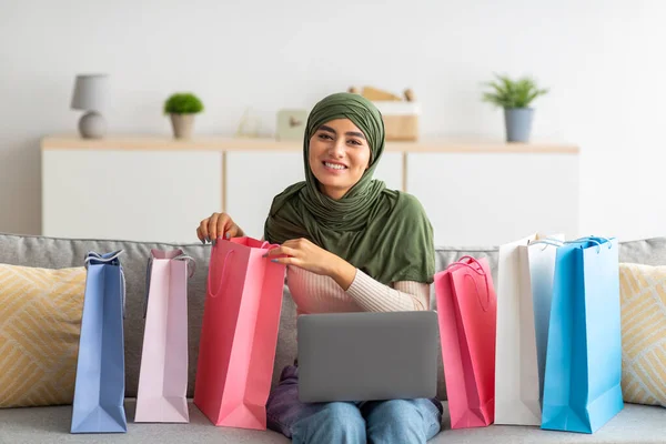 Hoş genç Arap bayan tesettürde oturuyor dizüstü bilgisayarla, parlak kağıt torbaların içine bakıyor. — Stok fotoğraf