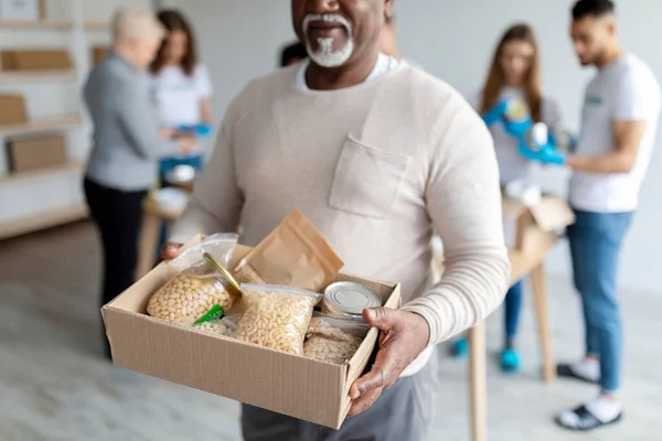 Ajuda humanitária. Africano americano homem idoso segurando caixa com donativos de alimentos, voluntários caixas de embalagem com produtos — Fotografia de Stock