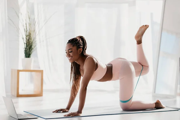 Young black woman working out her butt muscles in front of laptop, using elastic band, following video tutorial at home — Fotografia de Stock