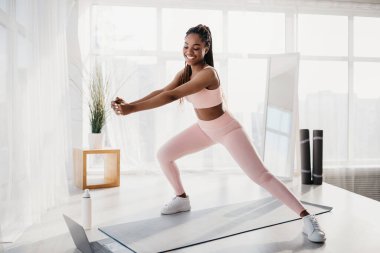 Çevrimiçi spor. Milenyum Afrikalı Amerikalı kadın paspas üzerinde egzersiz yapıyor, laptopta spor videoları izliyor, uzayı kopyalıyor.