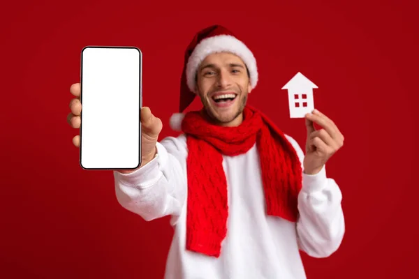 Aufgeregter Mann mit Weihnachtsmütze mit leerem Smartphone und Papierhaus-Ikone — Stockfoto