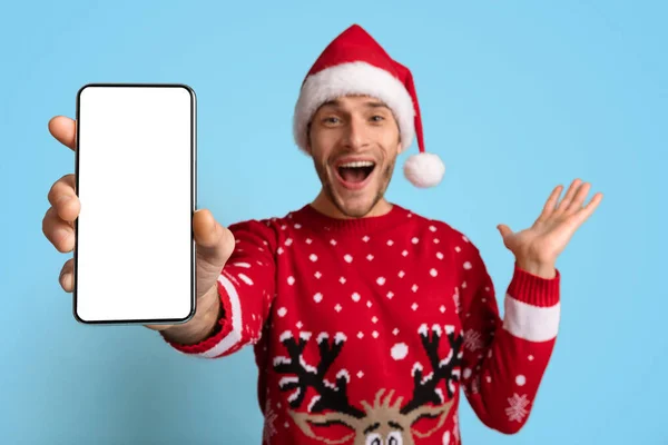 Urlaubswerbung. Aufgeregter Kerl mit Weihnachtsmütze demonstriert weißen Smartphone-Bildschirm — Stockfoto