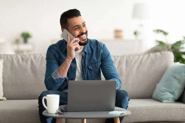 Gülümseyen genç bir Arap cep telefonuyla konuşuyor, laptop kullanıyor, evdeki koltukta oturuyor. — Stok fotoğraf