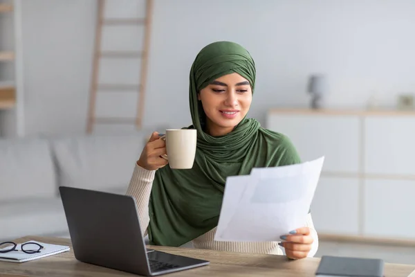 Arab hölgy hidzsábban kávét iszik, laptopot használ, üzleti dokumentumokat néz át az otthoni online munkaértekezlet során — Stock Fotó