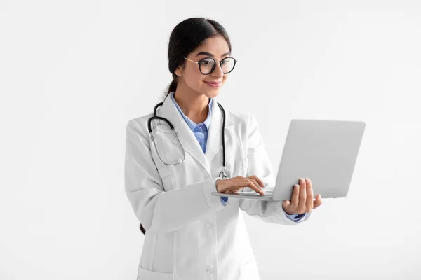 Busy millennial indian woman doctor in white coat and glasses working on laptop, isolated on white background — Stock Photo, Image