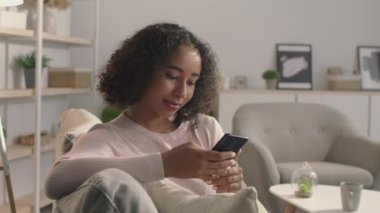 Çevrimiçi iletişim kavramı. Genç barışçıl Afro-Amerikan kadın internet sitesinde akıllı telefonuyla evde sörf yapıyor.