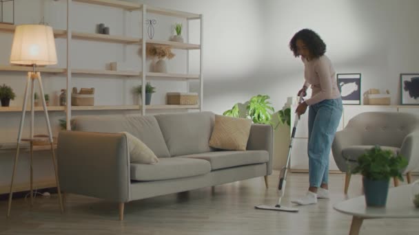 As tarefas domésticas. Jovem afro-americana lavando chão com esfregona, fazendo limpeza em casa na sala de estar, espaço livre — Vídeo de Stock